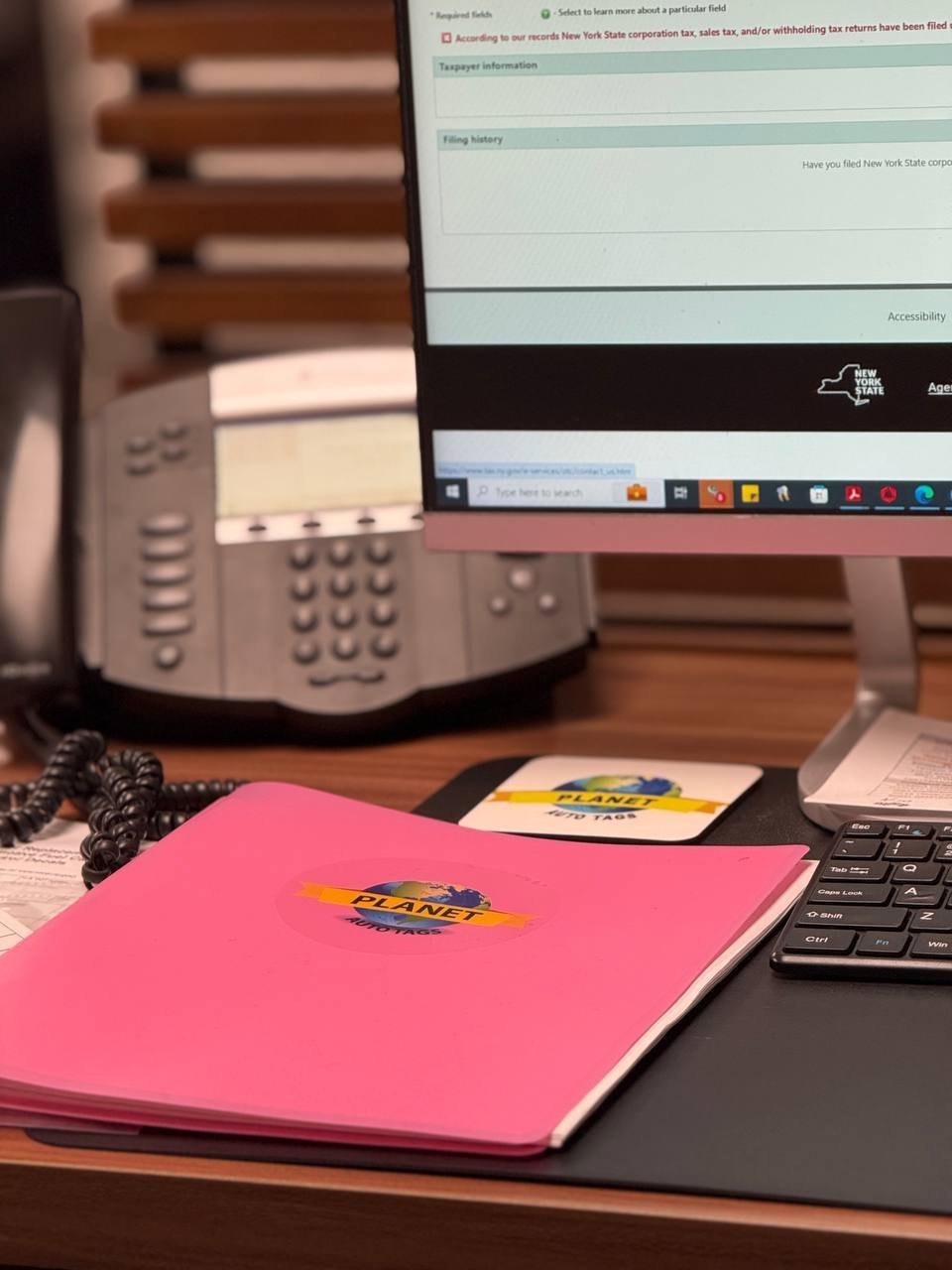 Office desk workspace with pink notebook, computer monitor, keyboard, phone, and office supplies on wooden surface